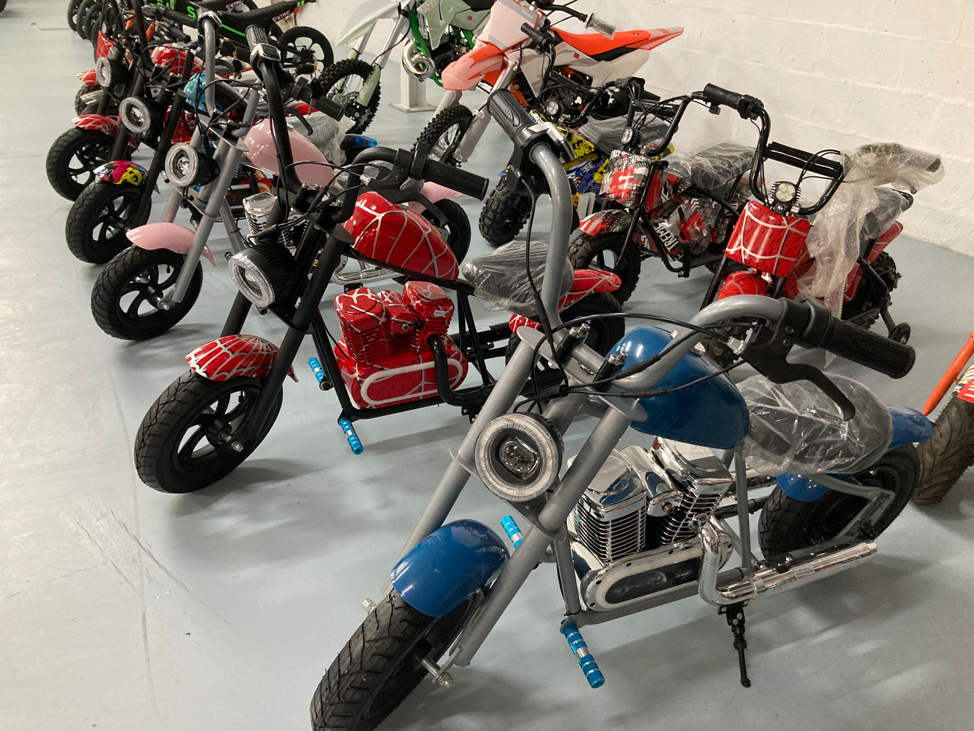 Minimotos eléctricas infantiles estilo chopper en gris con depósito azul metalizado, negra con telaraña roja y rosa claro, alineadas junto a pit bikes en la tienda