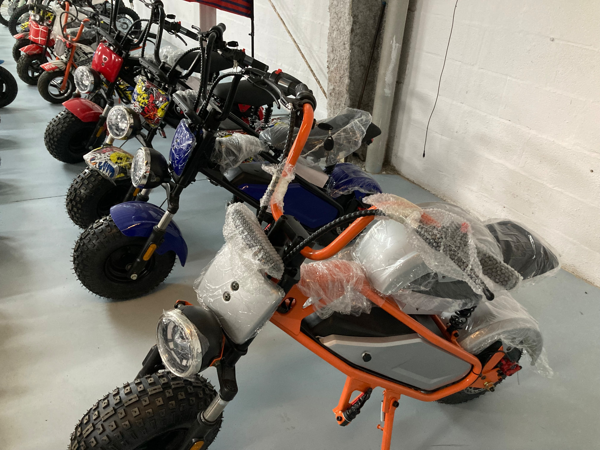 Minimotos eléctricas infantiles de ruedas anchas de tacos y faro redondo en naranja, azul, vinilo multicolor y rojo, protegidas con plástico de embalaje en la zona de montaje